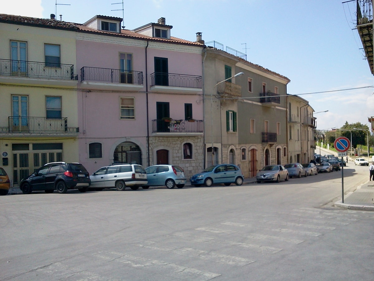 A street view of a charming town reveals a row of multi-story buildings with varying pastel colors. Balconies are seen on several floors, while parked cars line the cobblestone road. The setting includes a gentle slope leading to the distance, adding to the town's character.