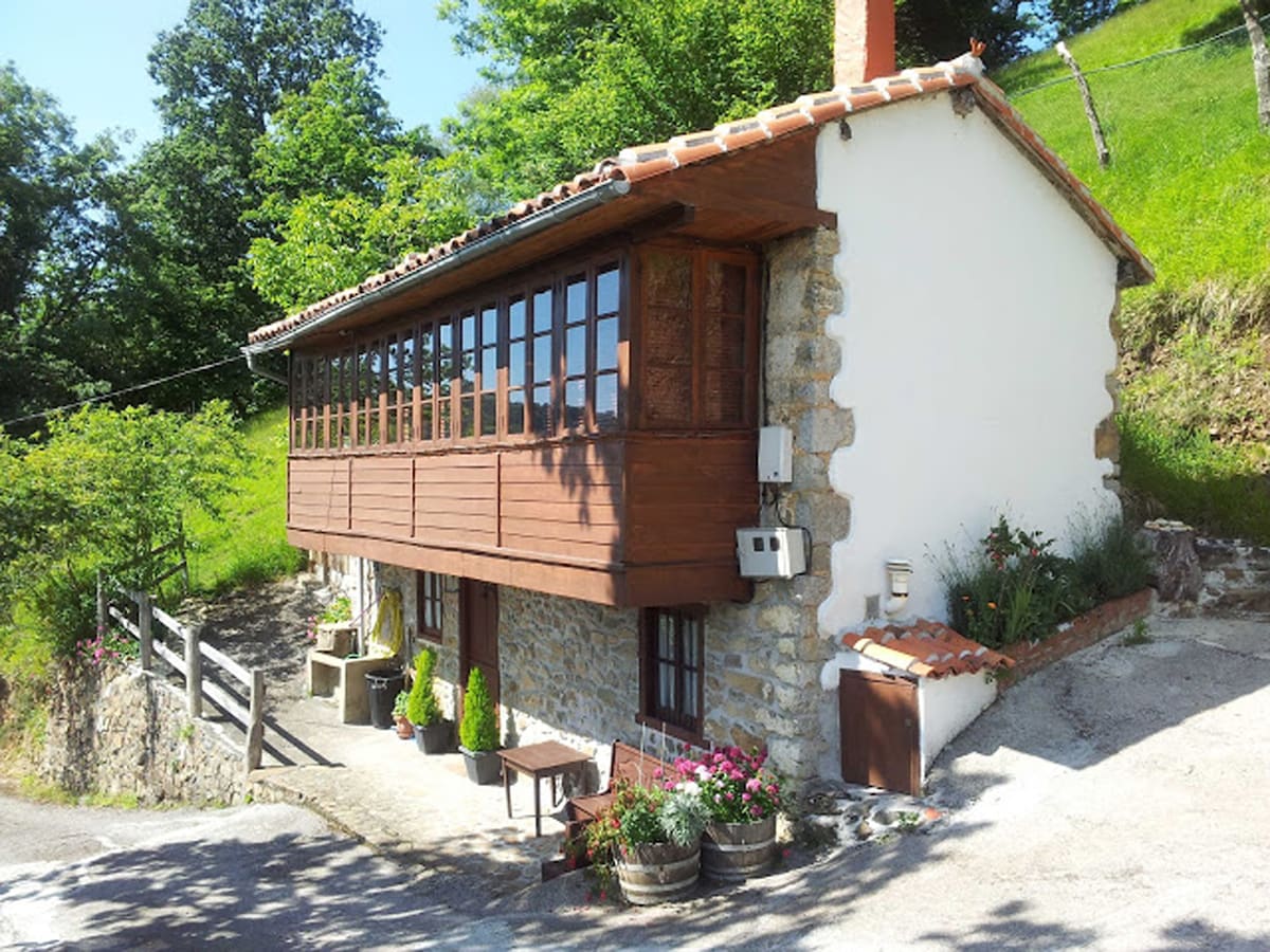 The exterior of a traditional Asturian house features a stone wall and wooden balcony. A tile roof and large windows provide a visual connection to the surrounding greenery. Potted plants and flower arrangements adorn the entrance, enhancing the home's rustic charm.