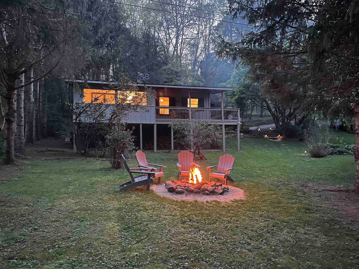 A fire pit is surrounded by adirondack chairs in a spacious yard, leading up to a two-story cabin nestled among trees. Soft light glows from the cabin windows, providing a warm ambiance as evening settles over the lush landscape.