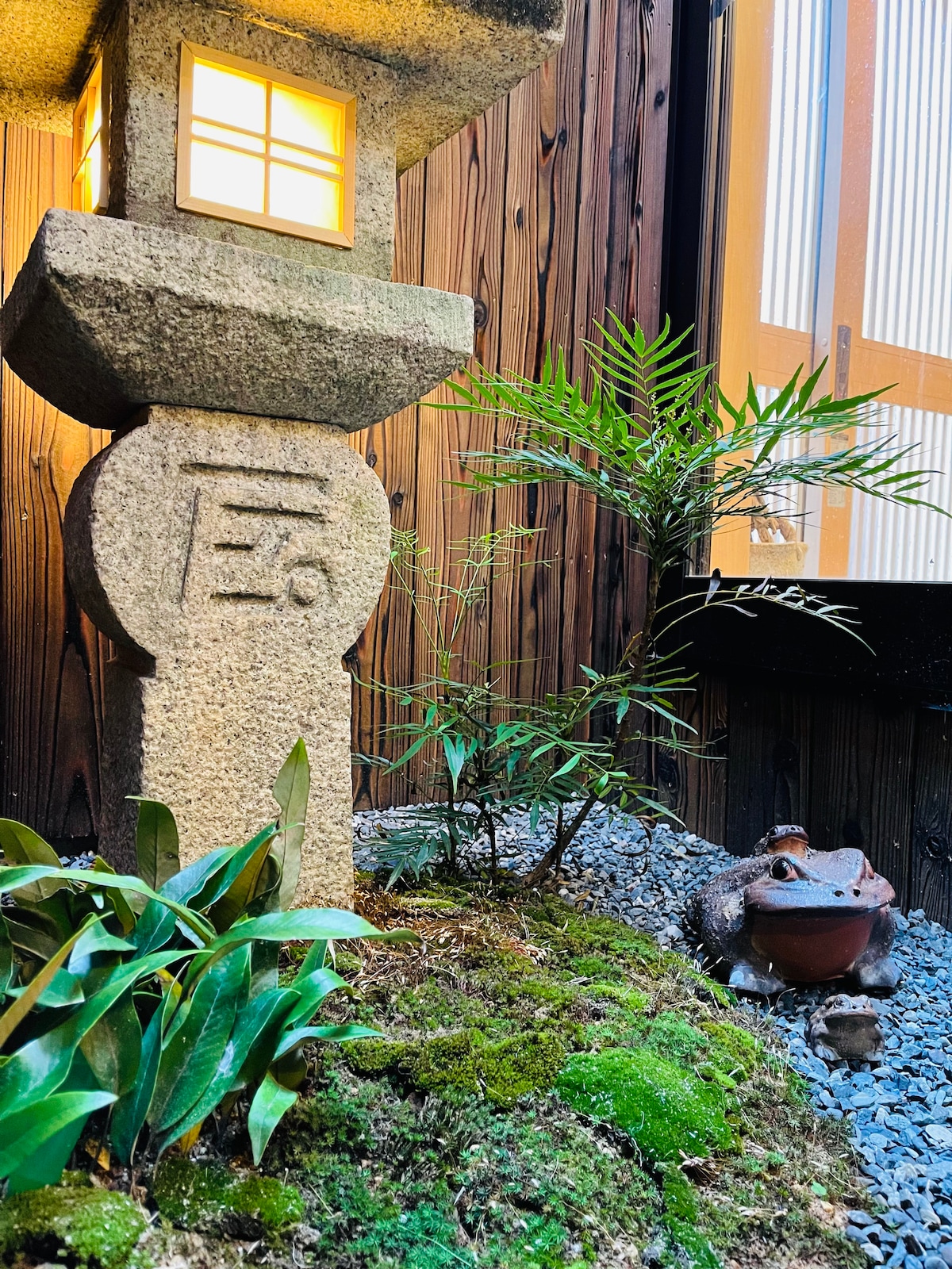 A traditional stone lantern illuminates a small garden area, surrounded by lush green plants and moss. Smooth stones cover the ground, and a ceramic frog figurine adds a charming detail, positioned near the plant life and lantern for added aesthetics.