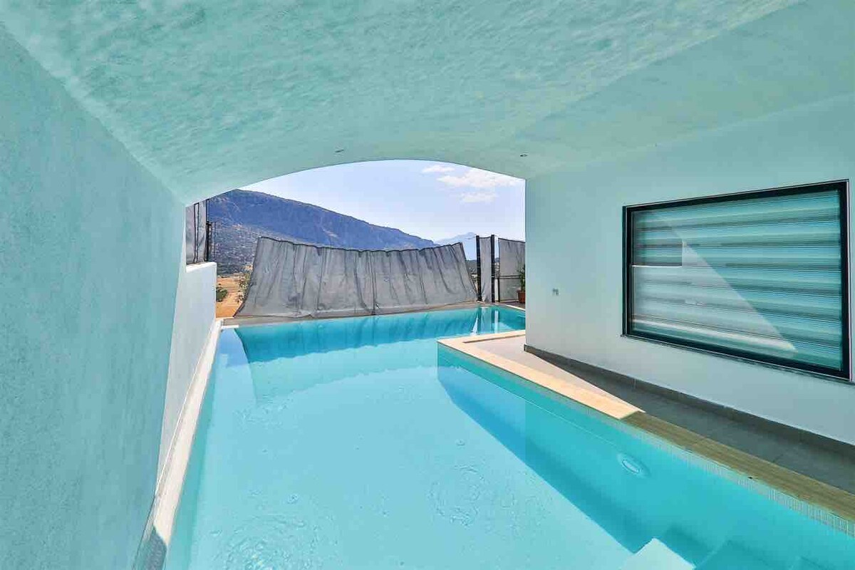 A private swimming pool is visible, featuring clear turquoise water. The pool area is enclosed by smooth white walls, with a view of the distant mountains. Sunlight illuminates the space, enhancing the serene atmosphere, while a window with horizontal blinds allows for natural light.