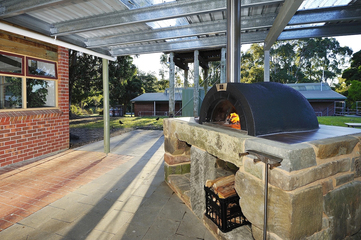 An outdoor space features a wood-fired pizza oven constructed from stone, with flames visible inside. The area is shaded by a metal roof and has stone flooring, blending with the natural surroundings. Windows of nearby buildings are visible in the background.