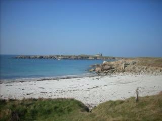 A sandy beach extends along the shoreline, meeting clear blue waters that reflect the sky. In the background, rugged rocks and a distant structure can be seen, framed by a tranquil landscape under a clear blue sky.