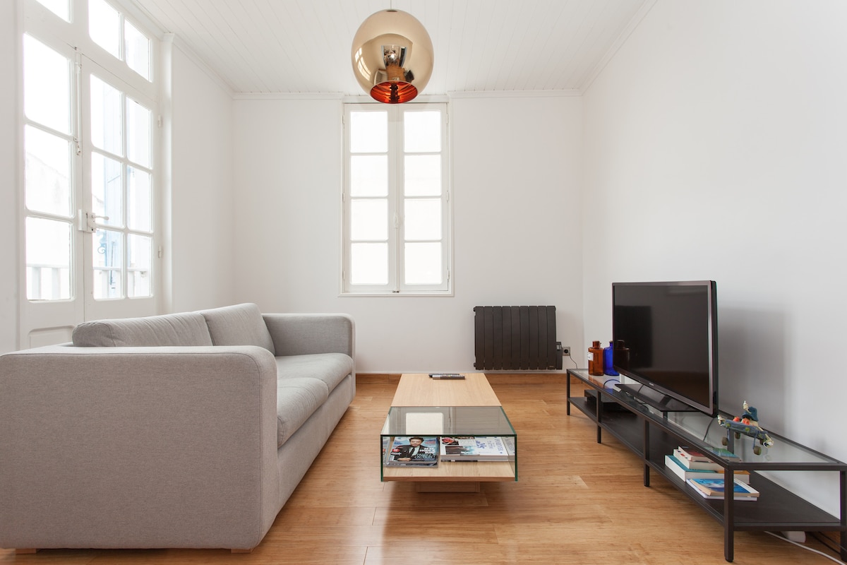 A bright living area is presented with a light-colored sofa facing a low glass coffee table and a TV on a sleek media unit. Large windows allow natural light to fill the space, complemented by a contemporary light fixture hanging above.
