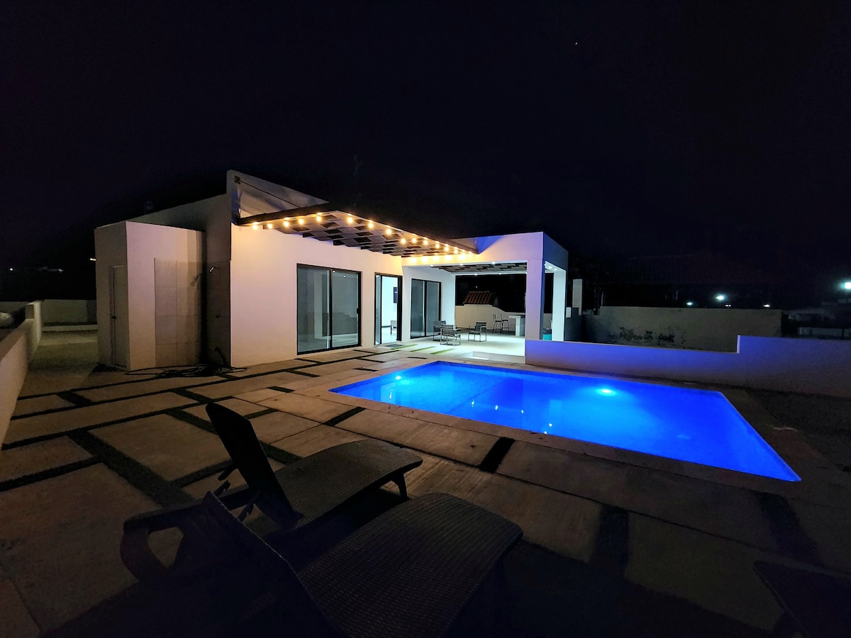 A modern outdoor area features a glowing blue pool surrounded by a spacious deck, illuminated by soft string lights. Large glass doors provide views of the interior, while lounging chairs offer a place to relax under the night sky.