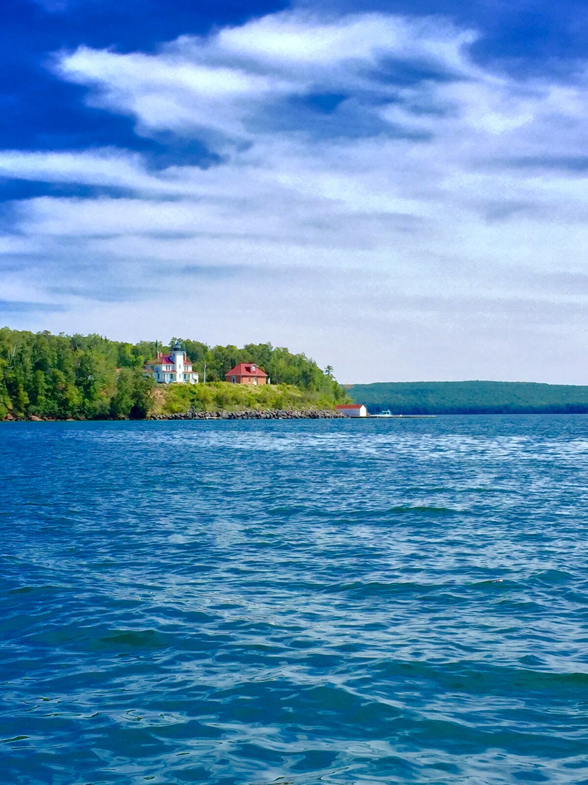 A scenic view of a lush, green shoreline is presented, with several cottages nestled among the trees. Calm blue waters reflect the sky, and gentle waves are visible in the foreground, enhancing the natural beauty of the secluded area.