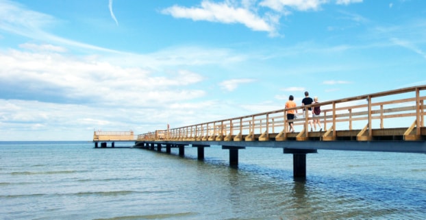 A wooden pier extends over the calm waters, supported by sturdy pillars. Two individuals can be seen walking along the pier, surrounded by the serene blue sky and gentle waves, contributing to a peaceful seaside ambiance.