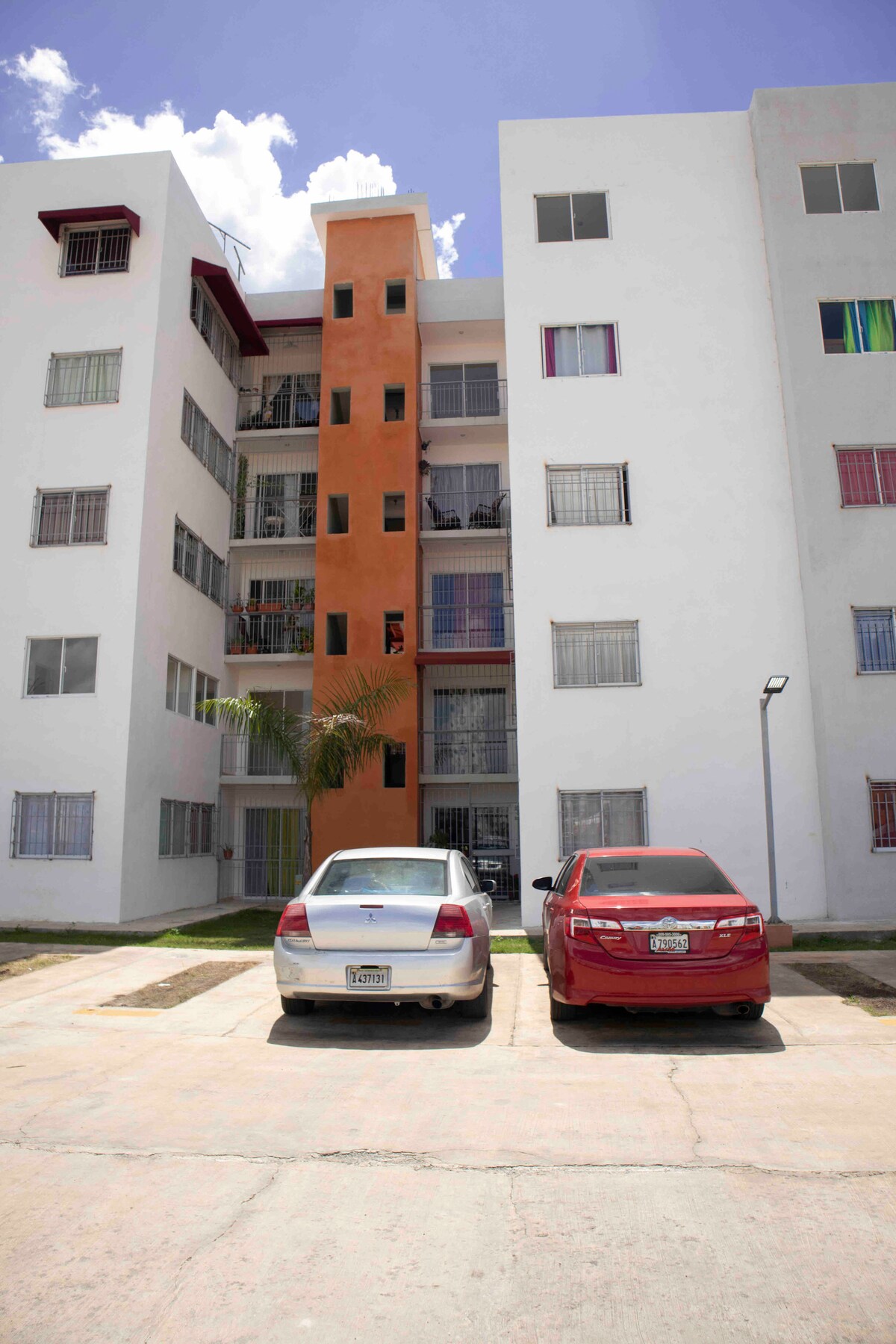 A multi-story apartment building is shown, featuring a mix of orange and white exterior walls. Two vehicles are parked in front, accompanied by a small palm tree and grassy areas. Several windows with light-colored curtains can be seen, providing an inviting facade.