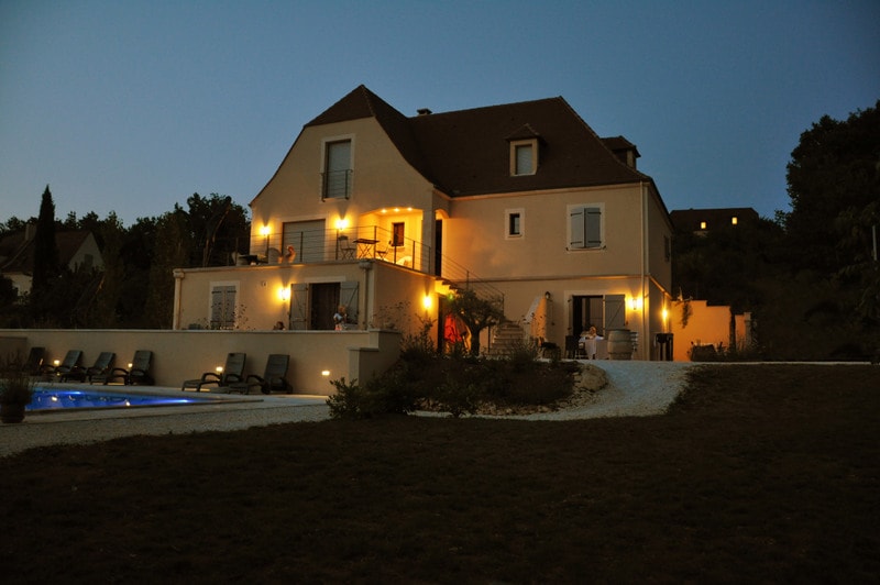 The spacious exterior of the Gite is highlighted at dusk, with warm lighting illuminating the building. A large terrace with seating overlooks the well-maintained lawn and nearby swimming pool, surrounded by nature. The tranquil atmosphere encourages relaxation in the quiet surroundings.