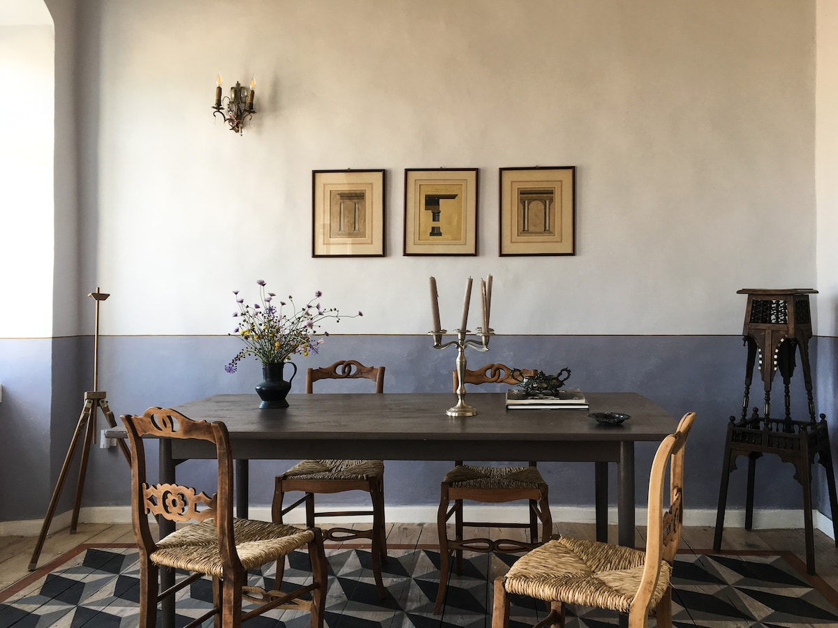 A dining area features a wooden table surrounded by four chairs, showcasing traditional craftsmanship. Framed artwork is displayed on the softly colored wall, above the table which is adorned with a vase of flowers. An elegant candle holder adds to the inviting setting.