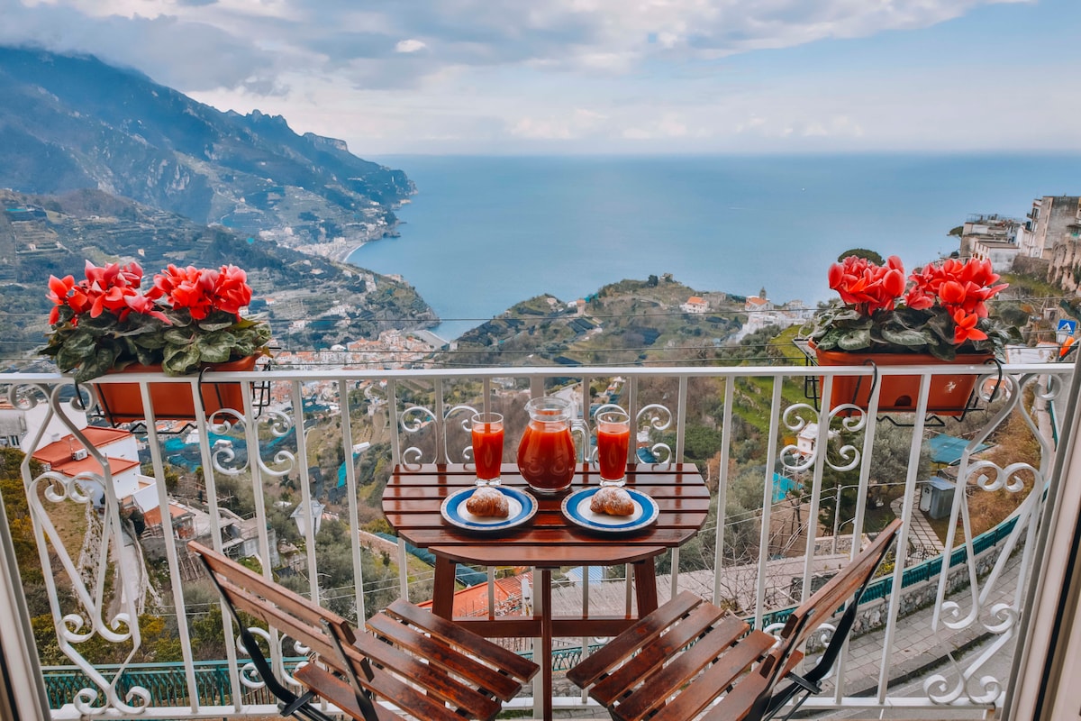 A balcony offering stunning views of the coastline features a small wooden table set with beverages and pastries. Surrounding flower pots add a splash of color, complementing the scenery of mountains and the sea in the background.