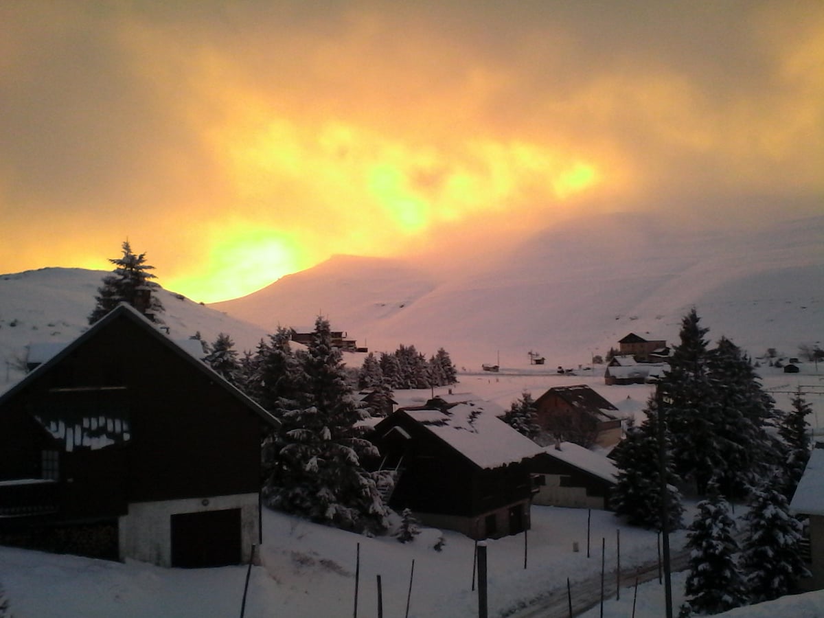 A serene winter landscape captures a sunlit horizon behind snow-covered mountains. The foreground features cozy chalets and trees blanketed in snow, with a pathway partially visible leading through the peaceful scene.