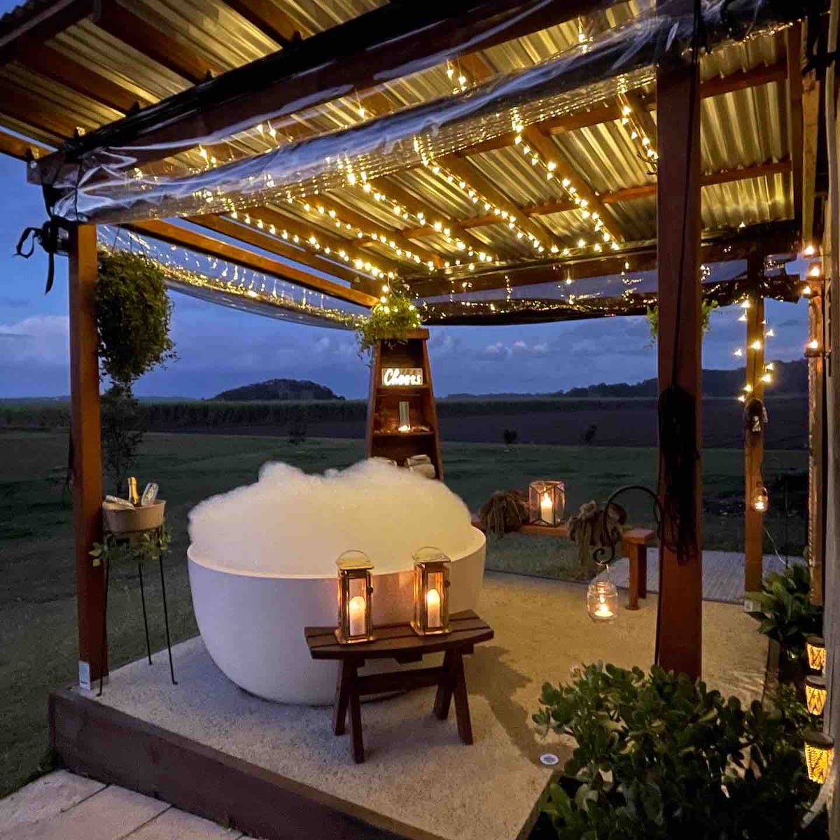 An outdoor bath area is adorned with string lights, creating a relaxing ambiance. A large white tub filled with bubbles sits on a stone surface, surrounded by lanterns and greenery. A tranquil view of the countryside is visible in the background.