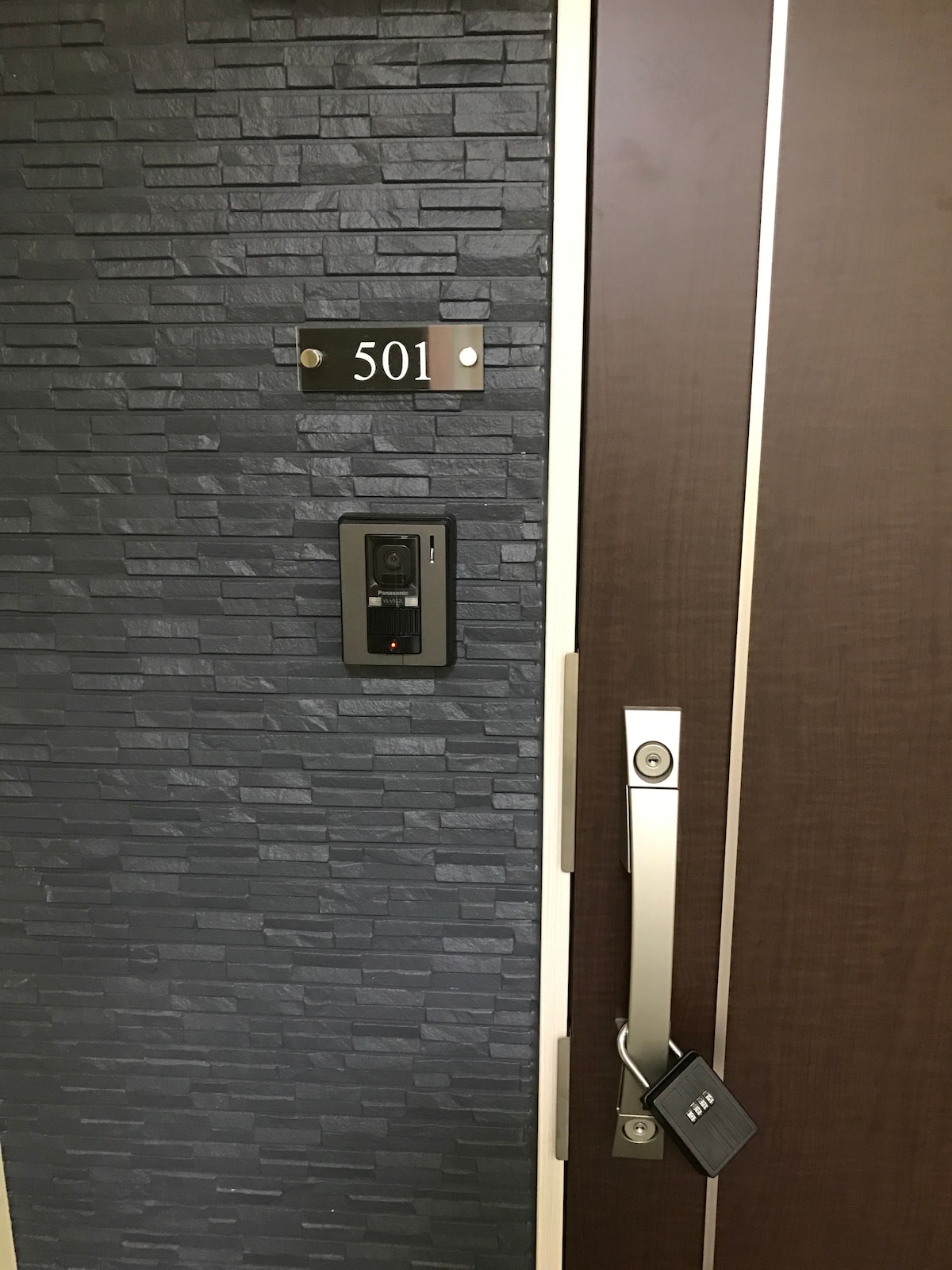 The exterior of the entrance door displays a dark stone wall complemented by a sleek wooden door. A nameplate indicating the room number '501' is mounted to the left. A modern intercom system is positioned beside the door lock, which features a secure padlock for added safety.