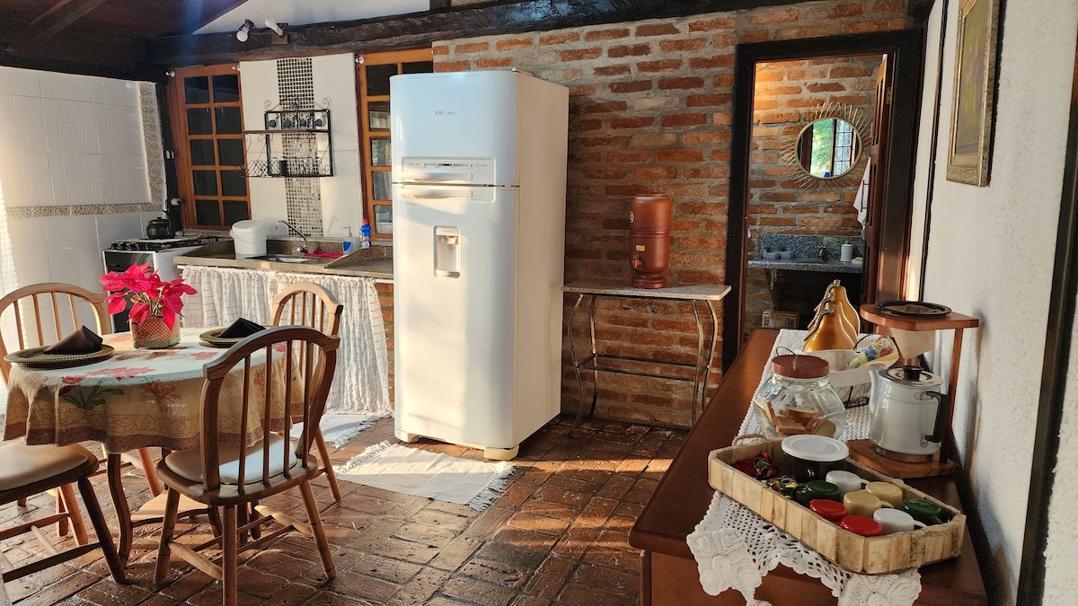 A rustic kitchen area is presented, featuring a wooden dining table set for four. A white refrigerator and a stone countertop complement the exposed brick walls. Natural light illuminates the space through the window, enhancing the inviting atmosphere of the room.