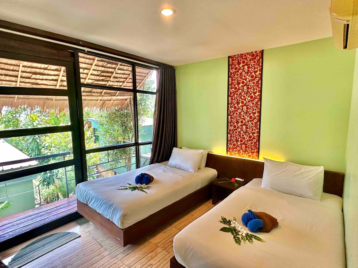 A bright and airy guest room is furnished with twin beds, each adorned with decorative towels and small plant accents. Large glass doors open to a private balcony, allowing natural light to fill the space, and greenery can be seen outside, enhancing the connection to nature.