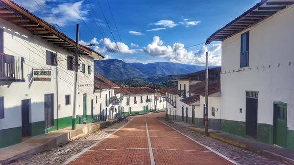 A serene view of a cobblestone street lined with white buildings featuring green accents. The road gently winds between the structures, leading towards the mountains in the distance under a clear blue sky. Bright sunlight casts soft shadows, enhancing the peaceful atmosphere.