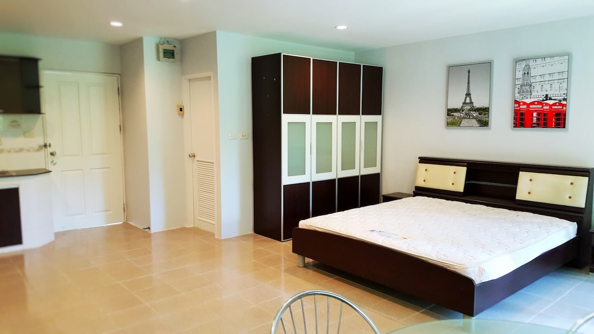 A modern studio space is shown with earthy tile flooring and a spacious bed positioned beside a large wardrobe containing glass panels. A dining table and chairs occupy one corner, while artwork depicting the Eiffel Tower and a red telephone box adorns the wall.