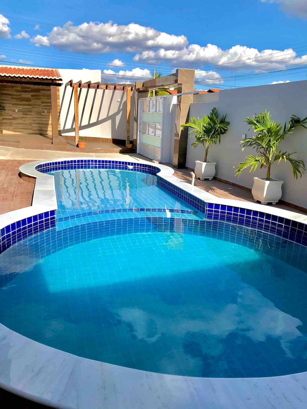 A sparkling outdoor pool is shaped creatively, surrounded by well-maintained landscaping. Bright blue tiles cover the pool interior, reflecting the sky above. White planters with greenery provide a fresh contrast against the smooth surface of the coping, creating a relaxing outdoor environment.