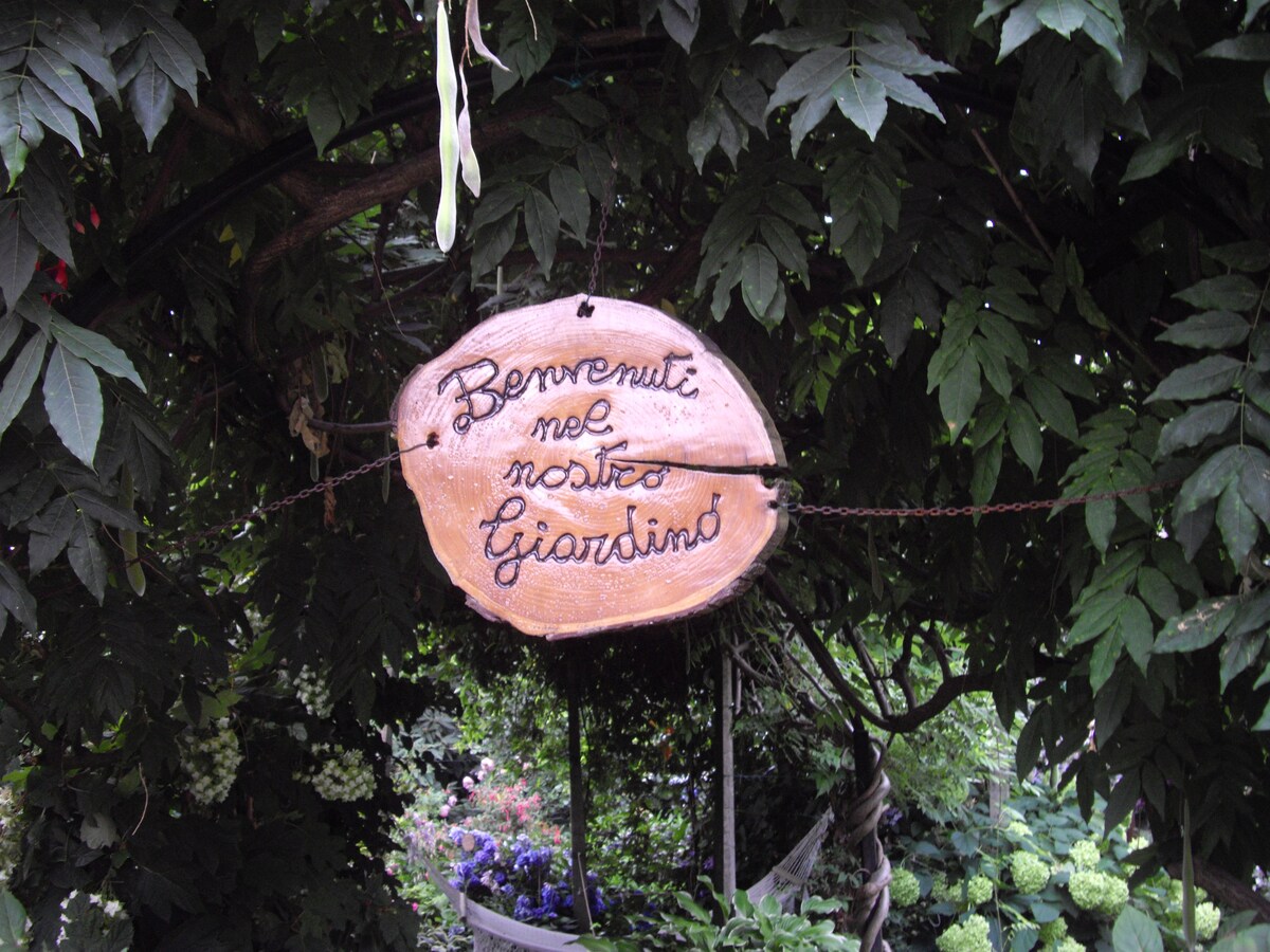 A wooden sign is hanging from a trellis, welcoming visitors to the garden. The sign's text is inscribed in a flowing script. Surrounding greenery provides a natural frame, enhancing the tranquil atmosphere of the space.