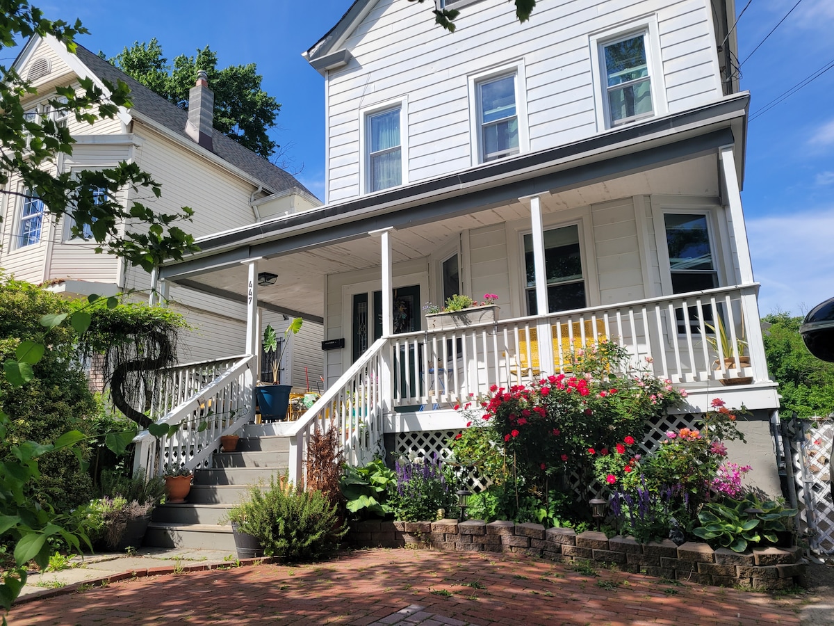 A charming front porch is framed by colorful flowers and greenery, creating a welcoming entrance. The house features white siding and large windows that allow natural light. A set of steps leads up to the porch, accompanied by various potted plants in a brick-paved area.