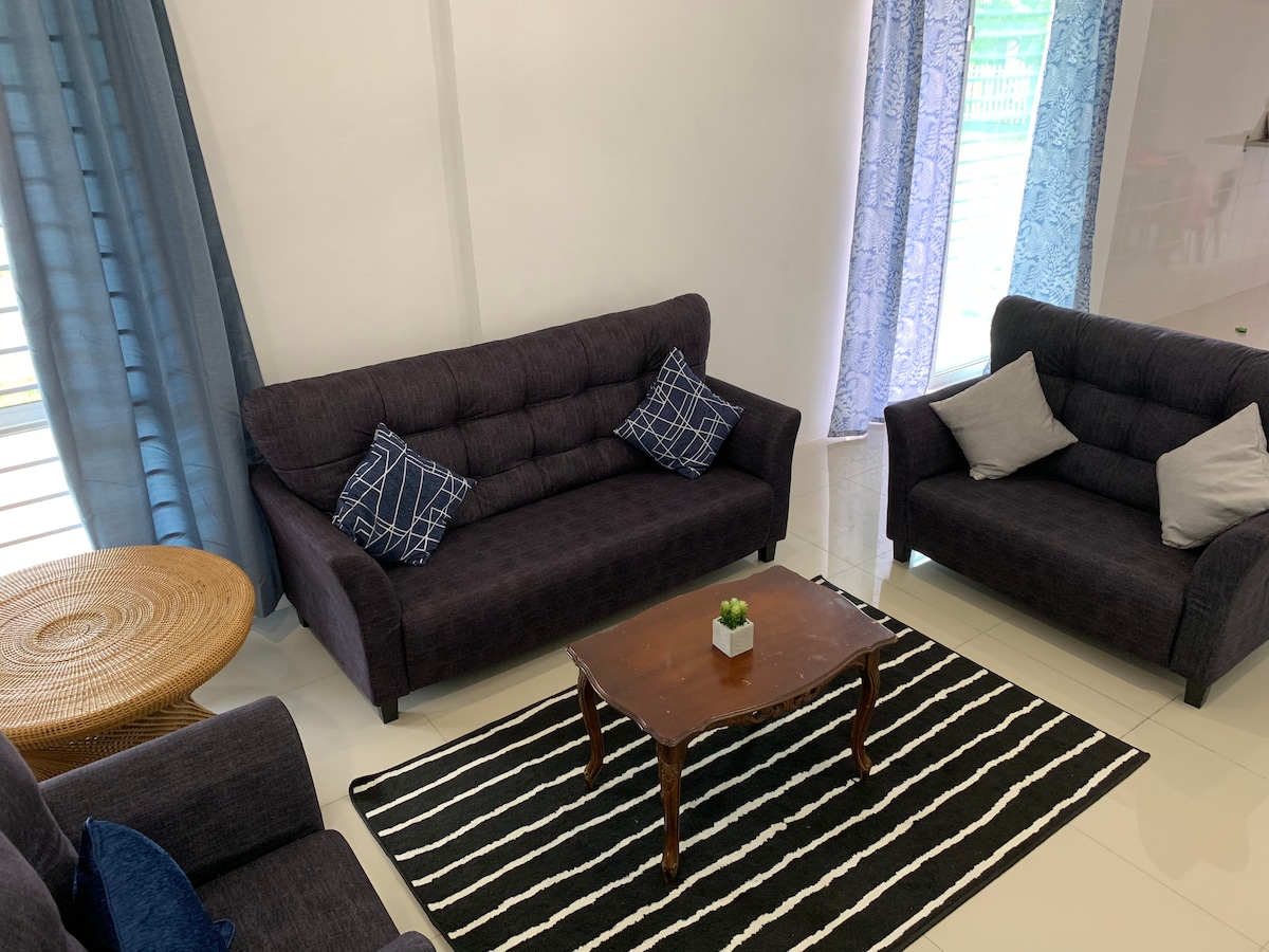 A cozy seating area is presented with two dark upholstered sofas, each adorned with decorative cushions. A circular wooden coffee table is positioned in the center, resting on a striped area rug. Natural light filters through sheer curtains, enhancing the space's ambiance.