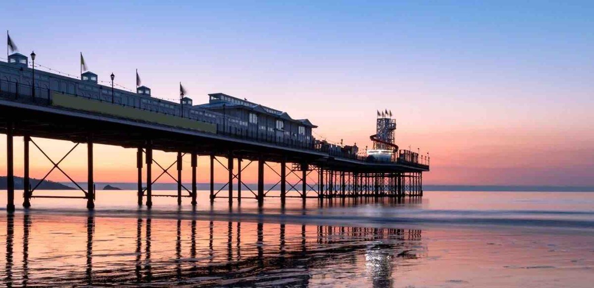 A seaside pier extends over calm waters, supported by tall wooden pillars. The sky transitions from soft orange to deep blue at sunset, reflecting colors on the water's surface. Flags can be seen fluttering along the pier, contributing to a serene coastal atmosphere.