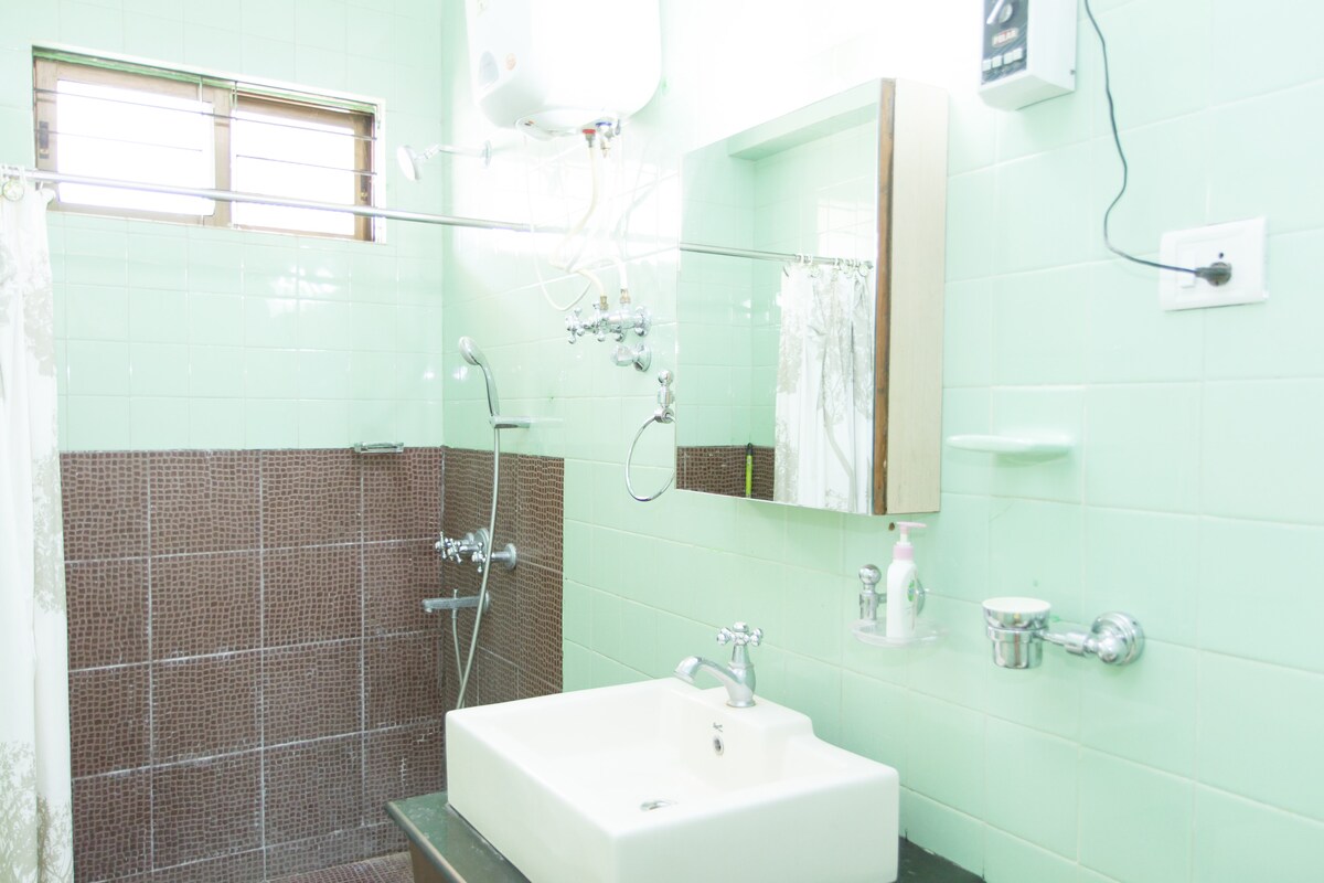 A clean, well-lit bathroom features mint green tiles with a contrasting brown tiled section. A modern sink with minimalistic fixtures is positioned on a dark countertop. A shower area is equipped with a wall-mounted faucet and a handheld showerhead, while a large mirror reflects the space.