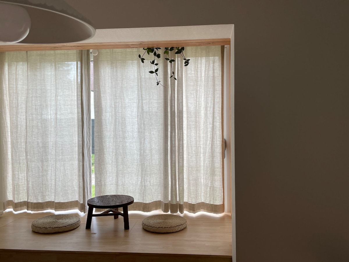A serene nook is framed by soft, light-colored curtains that gently filter natural light. Two round floor cushions are arranged around a low wooden table, creating a simple yet inviting space for relaxation or conversation.