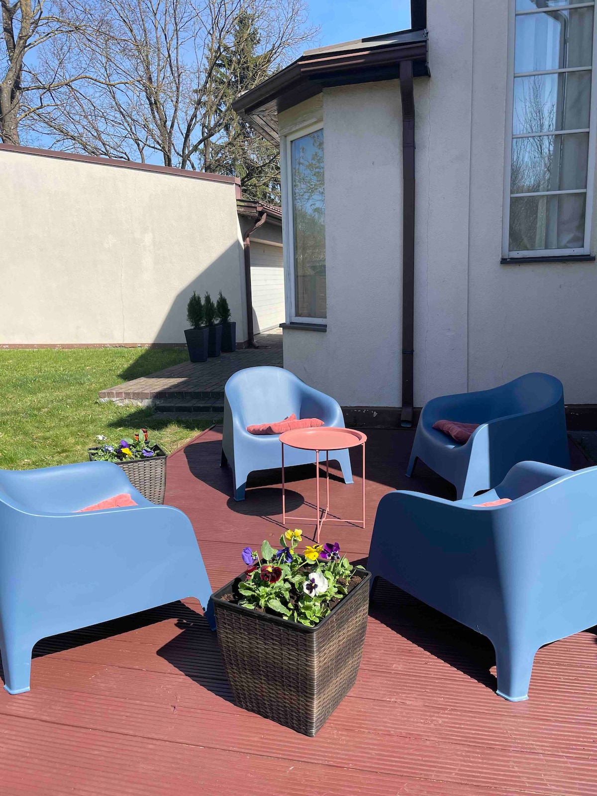 A spacious terrace features four blue armchairs arranged around a small pink table. A floral planter, filled with blooming plants, sits at the center, surrounded by a well-maintained lawn and light-colored exterior walls of the house.