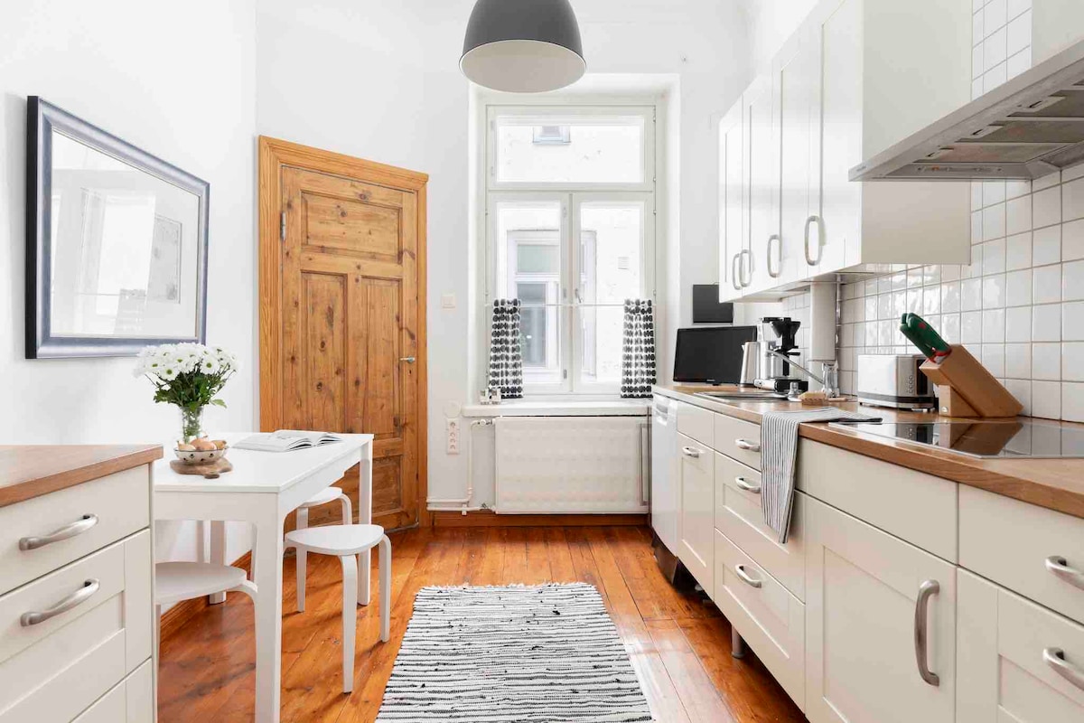 The kitchen features modern appliances and wooden countertop finishes. A small table with two chairs is positioned near the window, while a stylish rug lies on the polished wooden floor. Natural light illuminates the space, highlighting the minimalist design and cabinetry.