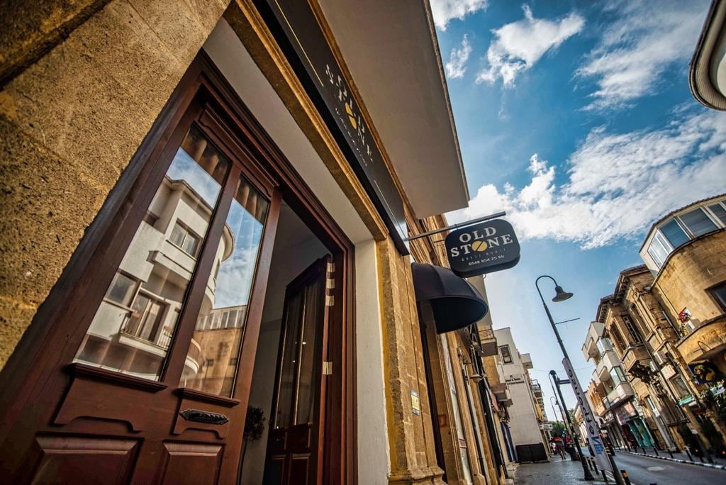 The exterior of a charming building is displayed, featuring a wooden door with detailed craftsmanship. Above, a sign reading 'OLD STONE' hangs, framed by a clear blue sky with scattered clouds. Stone accents highlight the architecture, blending with the surrounding urban landscape.