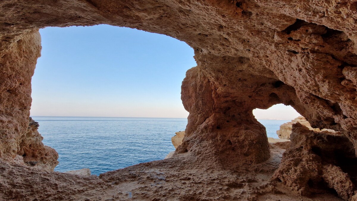 A natural rock formation creates two openings that frame the serene sea view. The calm waters reflect soft colors of the sky, with gentle waves visible in the distance. The rugged edges of the rocks offer contrast to the smooth horizon beyond.