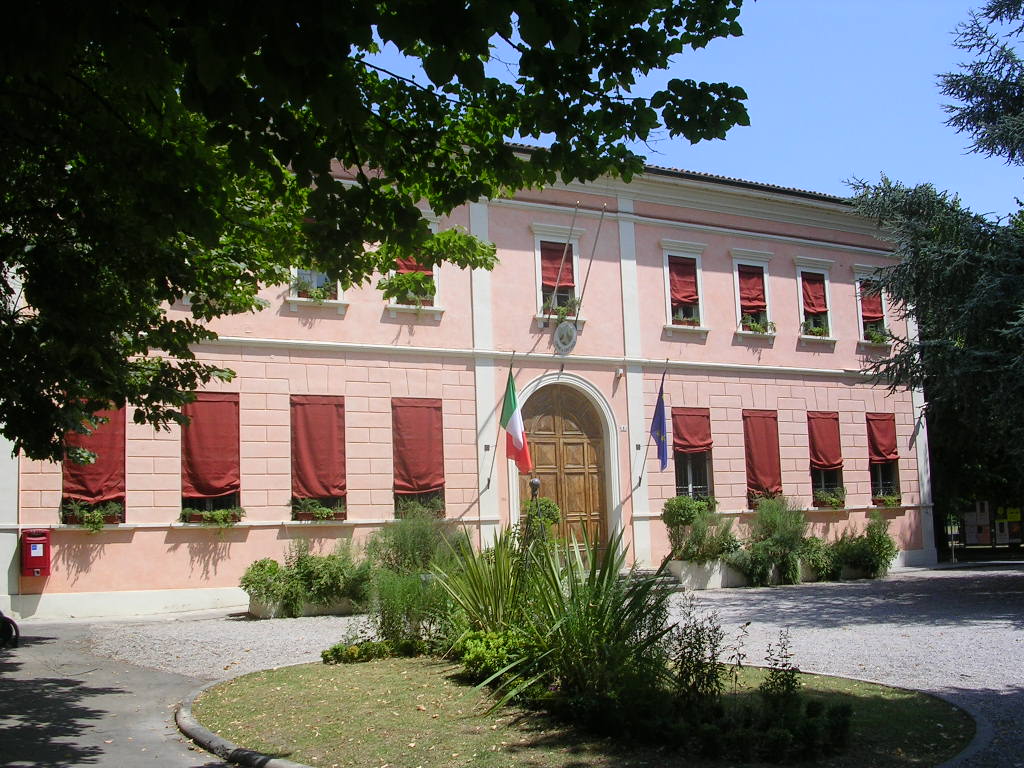 The building features a soft peach facade adorned with decorative elements. Red window coverings complement the greenery surrounding the entrance. A combination of national flags is displayed prominently in front, enhancing the sense of place.