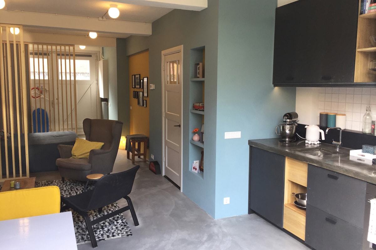 A studio space features an open layout with a living area and kitchenette. A comfortable armchair and a small table are visible. The kitchen includes modern appliances and wooden cabinets, while decorative items are arranged on shelves. Natural light brightens the room from nearby windows.