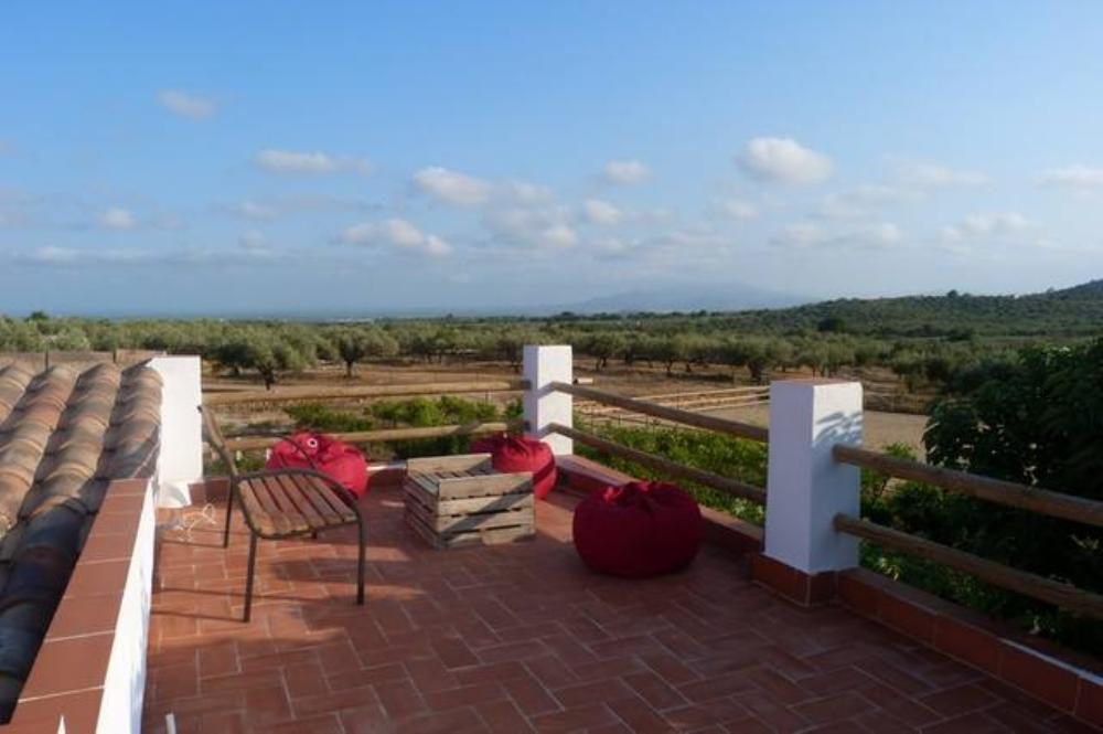 A rooftop terrace provides a relaxing setting with views of the surrounding olive groves and distant hills. Comfortable bean bags and a small wooden table are placed on the terracotta tiled floor, complemented by a single metal chair. The sky is bright with scattered clouds.