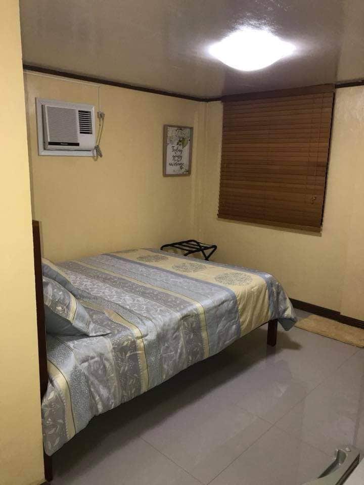 A compact room features a spacious bed with a striped bedspread and a wall-mounted air conditioning unit. Natural light enters through a window adorned with wooden blinds. A small folding chair is positioned near the bed, complementing the neutral-colored walls and tiled floor.