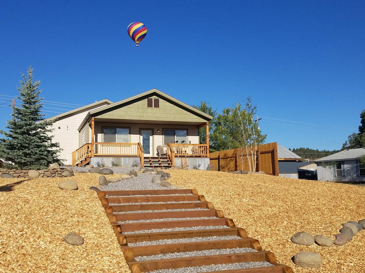 The exterior of a newly built house is showcased, featuring a light green façade and a welcoming front porch. A pathway made of wooden steps leads up the gently sloped yard, accented by gravel and decorative stones. A colorful hot air balloon floats in the clear blue sky.