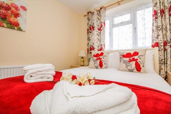 A bright room features a spacious bed dressed in white linens and adorned with decorative pillows. A vibrant red throw blanket adds warmth. Nearby, a tasteful floral painting complements the curtains, and a bedside lamp provides soft lighting.