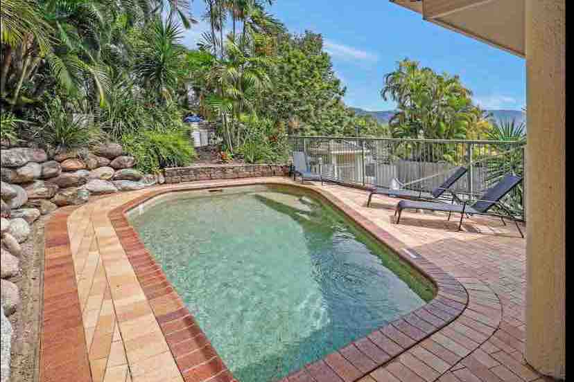 A private pool is framed by lush tropical landscaping, with a surrounding deck featuring two lounge chairs positioned for relaxation. The pool's clear water reflects surrounding greenery under a bright sky, creating a serene outdoor space.