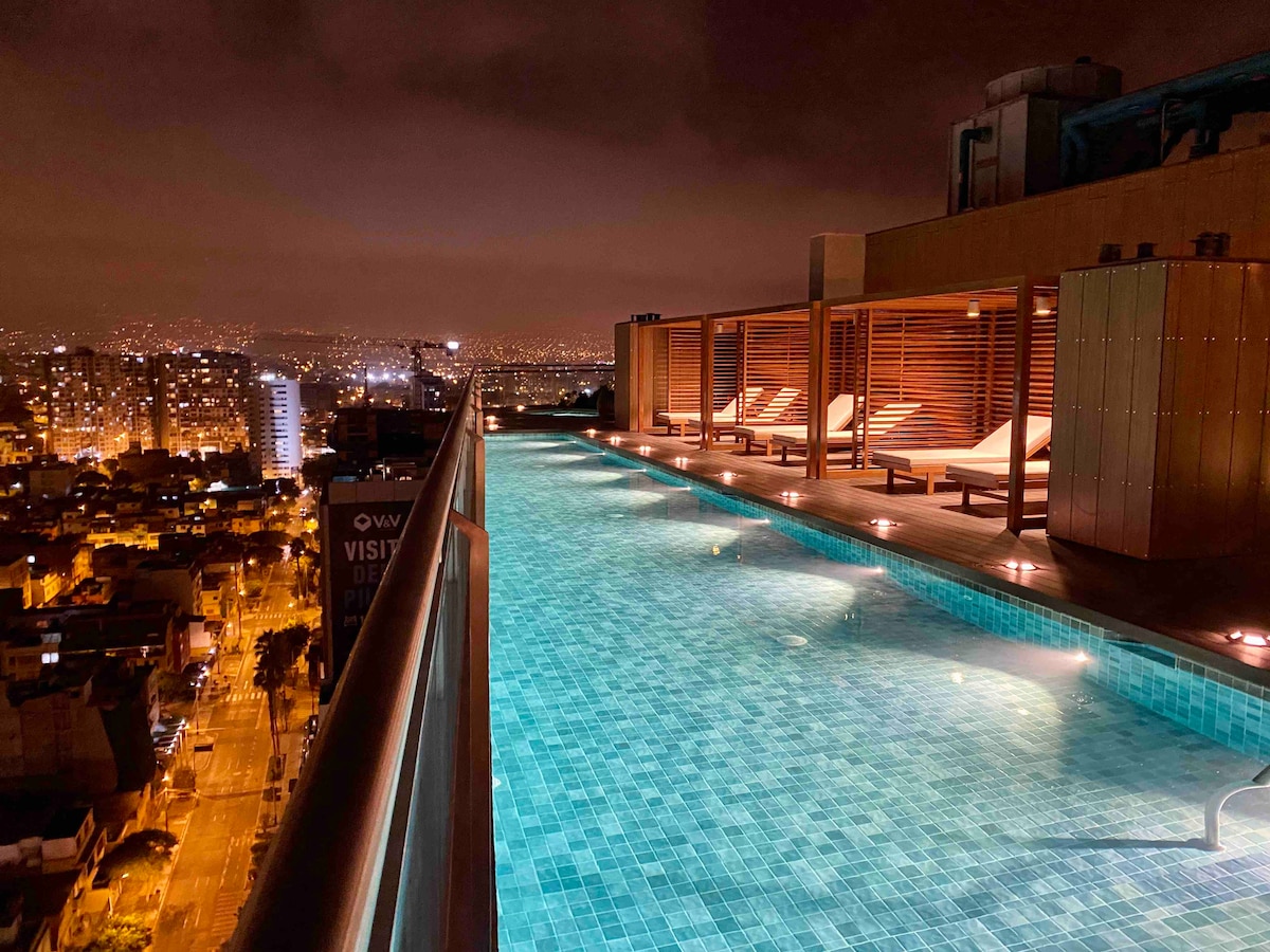 An illuminated rooftop pool offers panoramic views of the city skyline at night. Soft lighting reflects off the water's surface, and sun loungers are positioned along the edge, providing a serene space for relaxation.