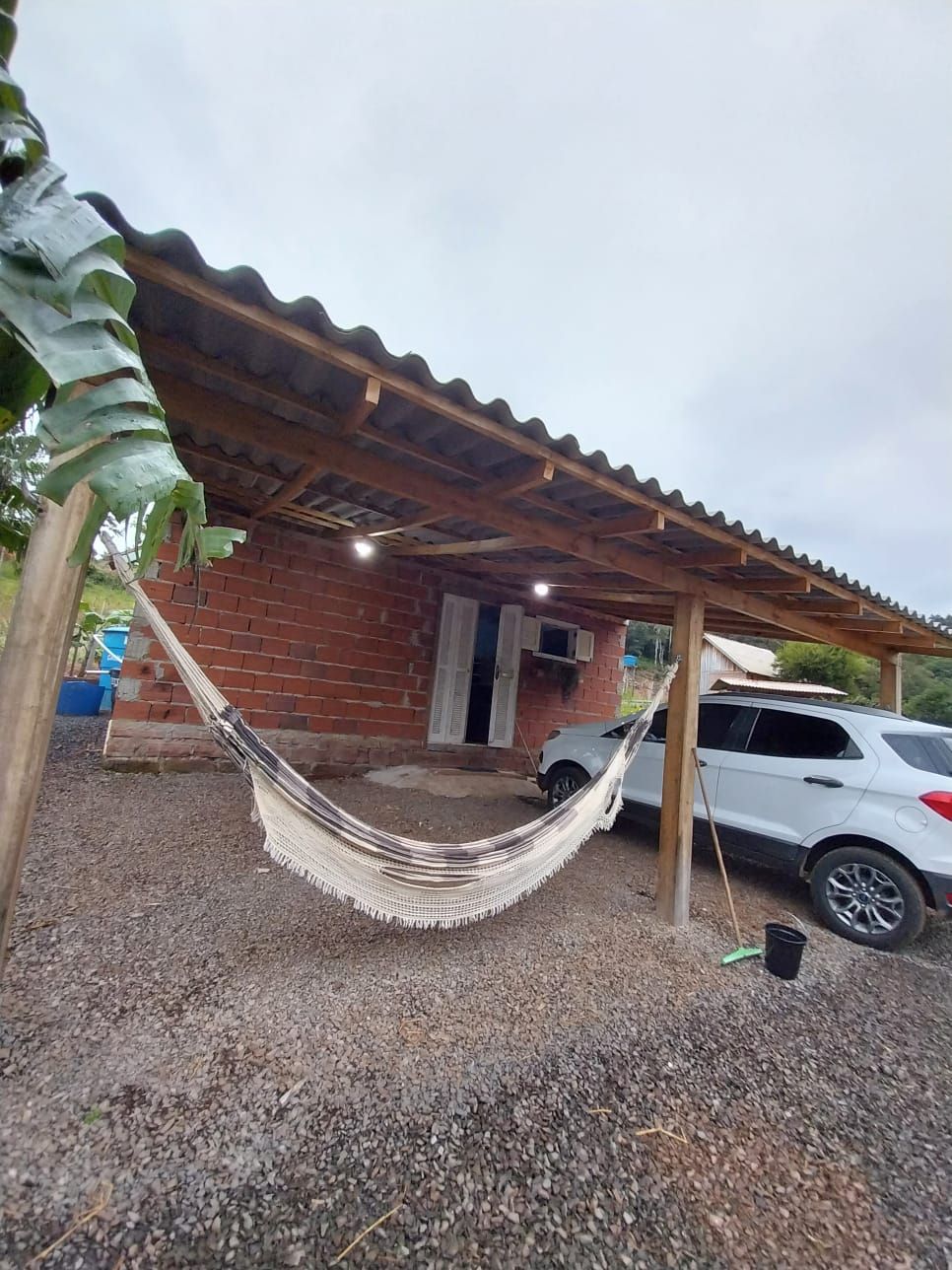 A cozy outdoor area is featured, with a sturdy hammock suspended under a shaded wooden structure. A white vehicle is parked nearby on a gravel surface, and simple lighting enhances the space's functionality. The brick building in the background provides a rustic charm.