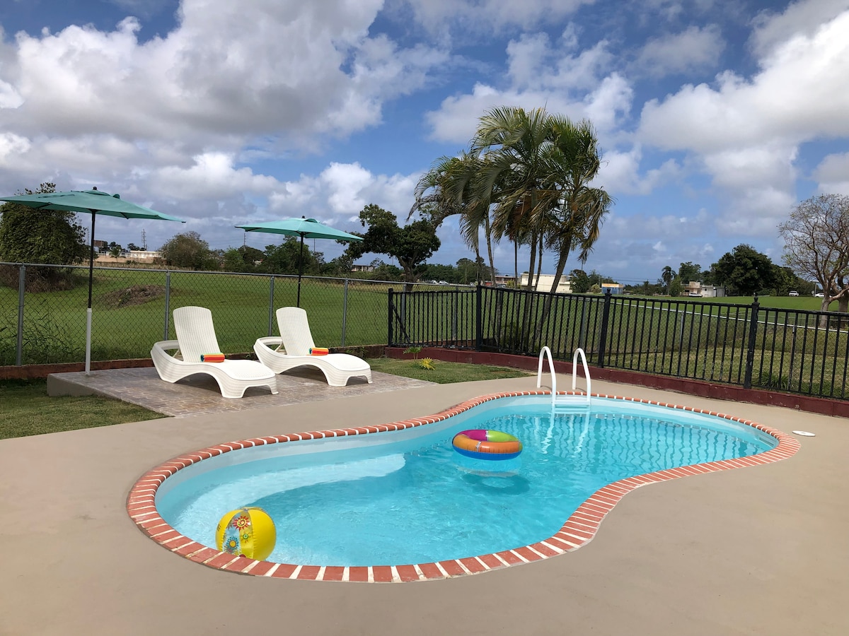 A private pool area features an inviting blue pool surrounded by a spacious concrete deck. Two lounge chairs are positioned under teal umbrellas, and colorful inflatable pool toys float on the water. A fenced lawn with greenery is visible in the background.