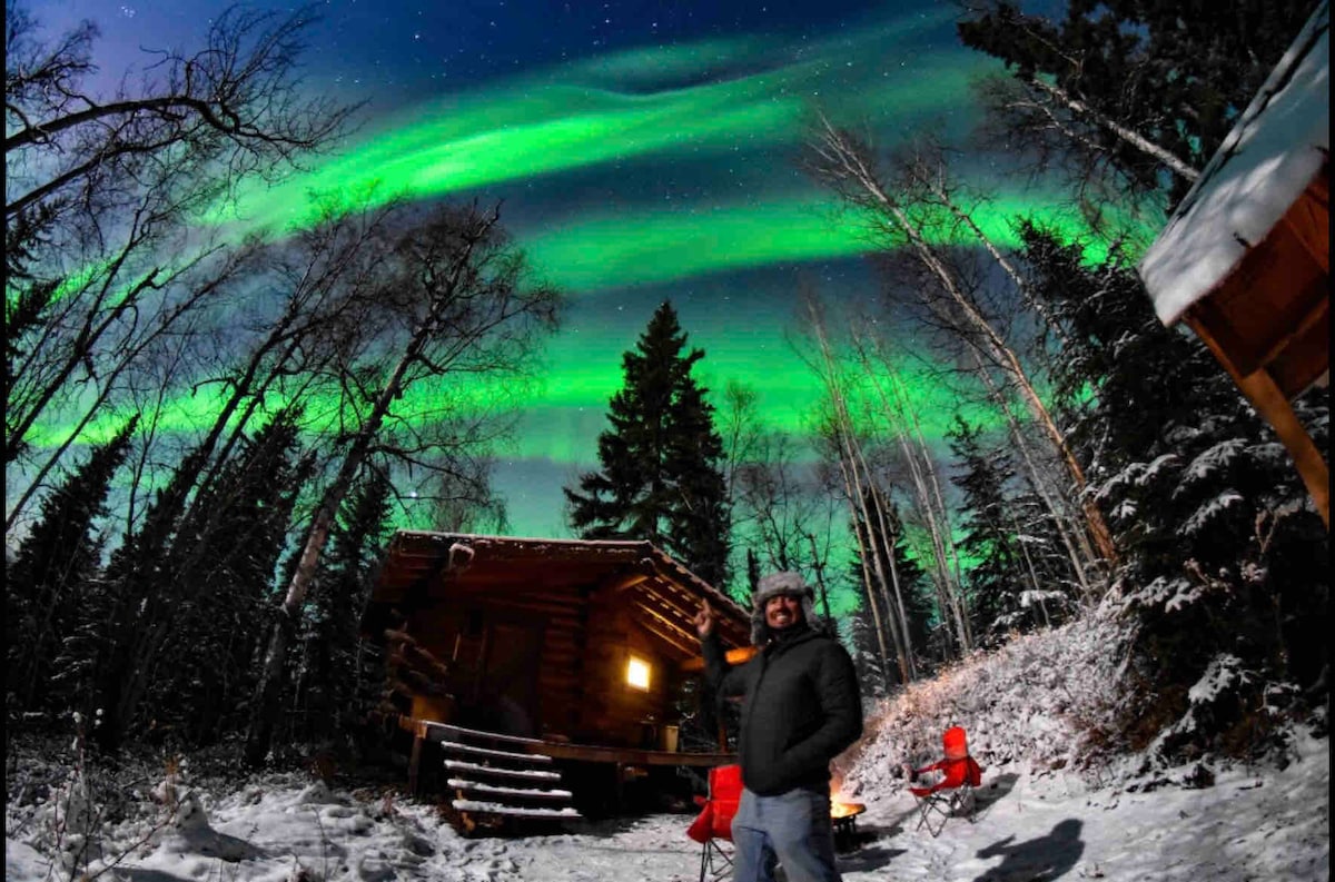 The image captures a cabin surrounded by snow-covered trees, illuminated by vibrant green auroras dancing in the night sky. Two red chairs are positioned on the ground nearby, with a figure standing beside them, looking upwards toward the captivating display above.