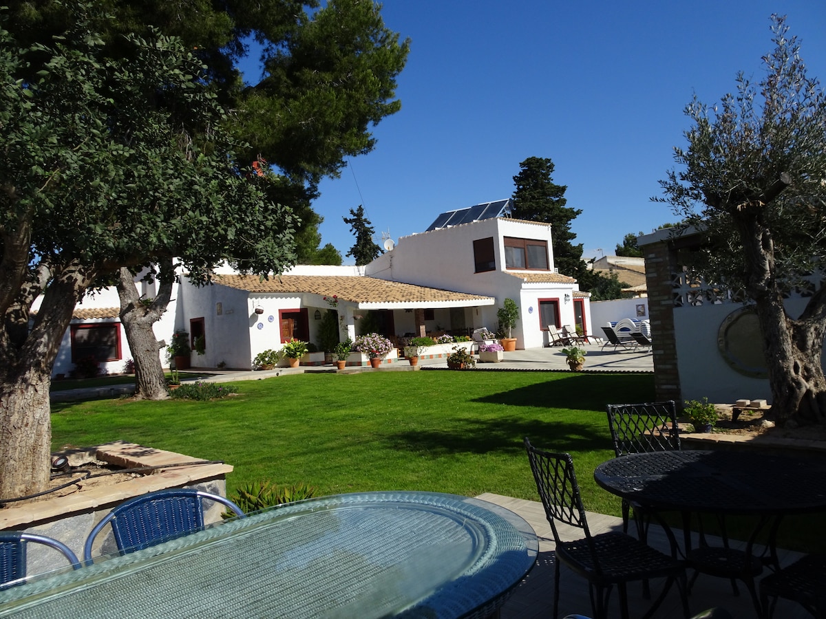 A rustic villa is set amidst a spacious garden, adorned with greenery and well-maintained landscaping. Multiple seating areas are visible on the stone patio, featuring metal tables and chairs. The villa's exterior showcases large windows and a tiled roof, with sunloungers positioned for relaxation.