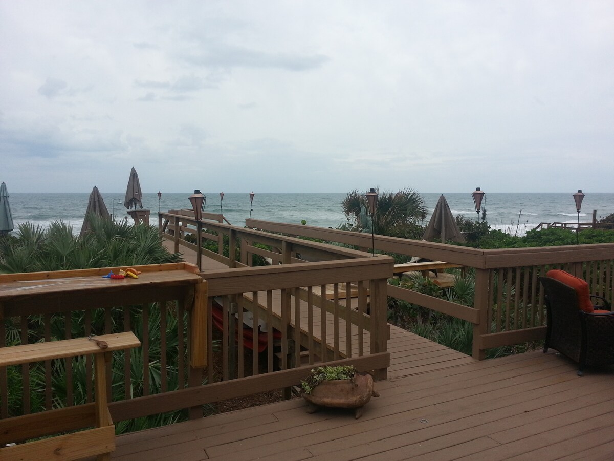 A large outdoor deck overlooks the ocean, featuring natural wood railings and comfortable seating areas. Umbrellas provide shade, while palm leaves and other coastal vegetation frame the view. The sound of waves adds to the serene atmosphere, evoking a tranquil beach experience.