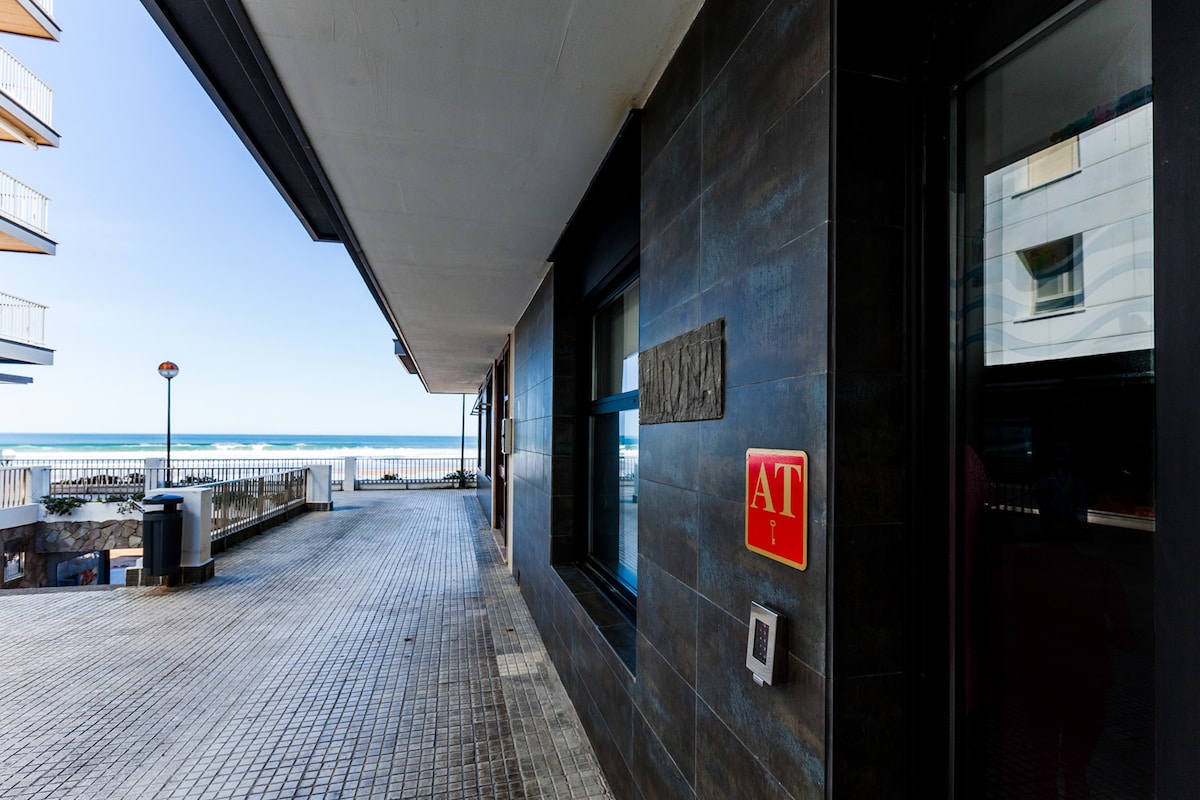 A coastal view is offered from the entrance of the apartment, showcasing the ocean beyond. The modern building features dark exterior finishes, and a spacious walkway is visible, with nearby amenities, including a trash bin and street signs.