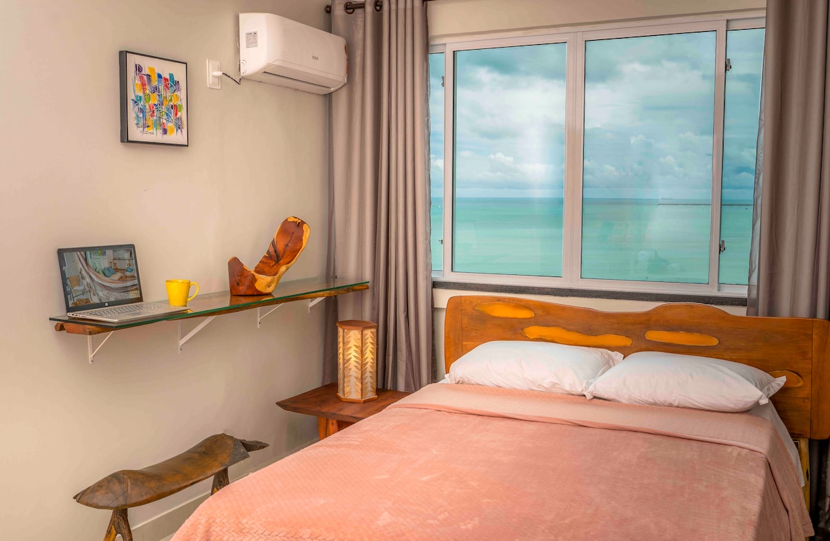 A cozy bedroom features a spacious bed with light bedding and contrasting yellow accents. A wooden shelf holds a laptop and a yellow cup, complemented by an artistic piece on the wall. A large window offers a view of the water, with natural light flowing through.