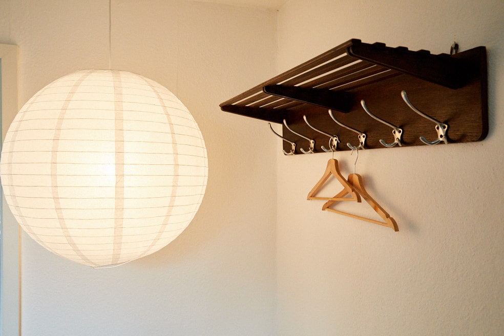 A softly illuminated round paper lamp hangs from the ceiling, casting a gentle glow. A wooden coat rack with multiple hooks is mounted on the wall, featuring three wooden hangers, providing a functional and organized space.