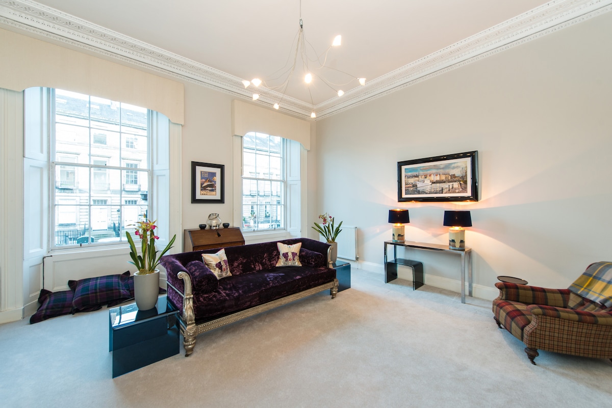 The living area features large windows allowing natural light to fill the space. A plush purple sofa with decorative pillows is positioned against the wall, complemented by a patterned armchair. A stylish console table, with lamps, adds functionality to the room.